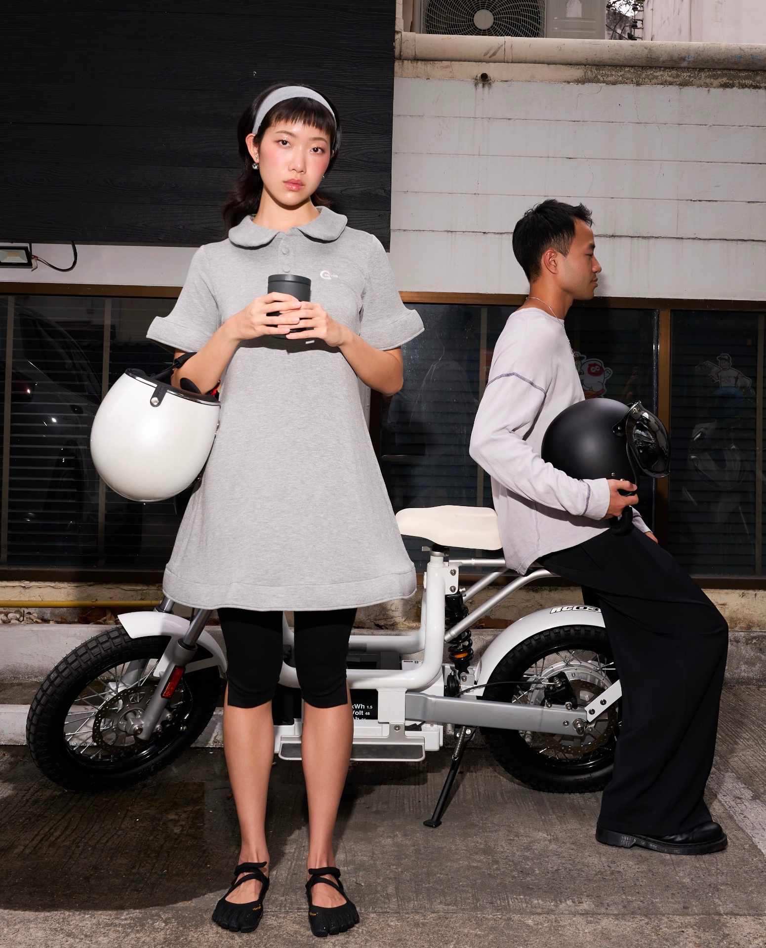 Two models with HELMYY helmets beside a white electric bike — matte black helmet with tinted shield and pearl white helmet with clear shield